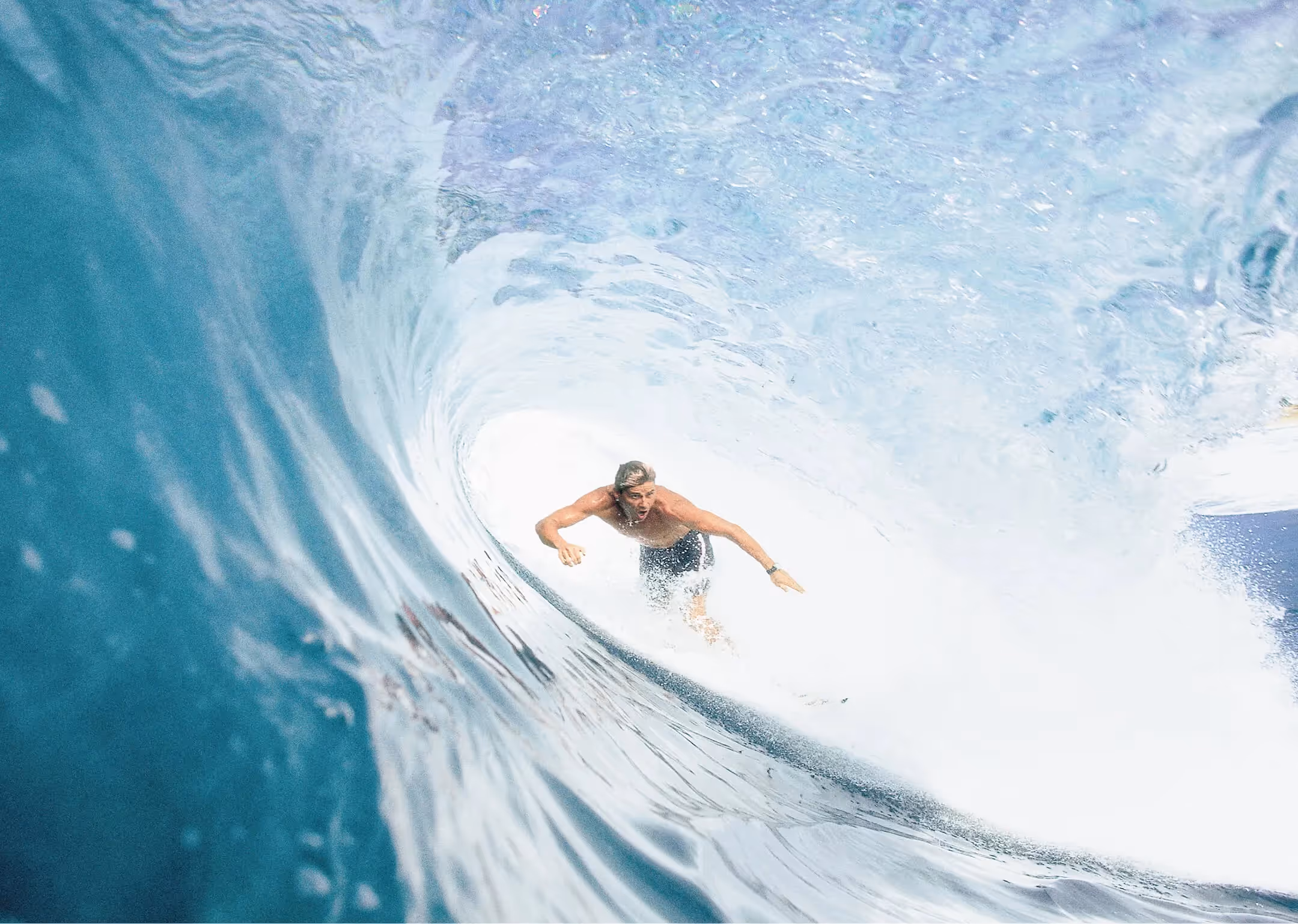 Surfer reitet tief in der Röhre einer grossen türkisfarbenen Welle bei hellem Tageslicht.

