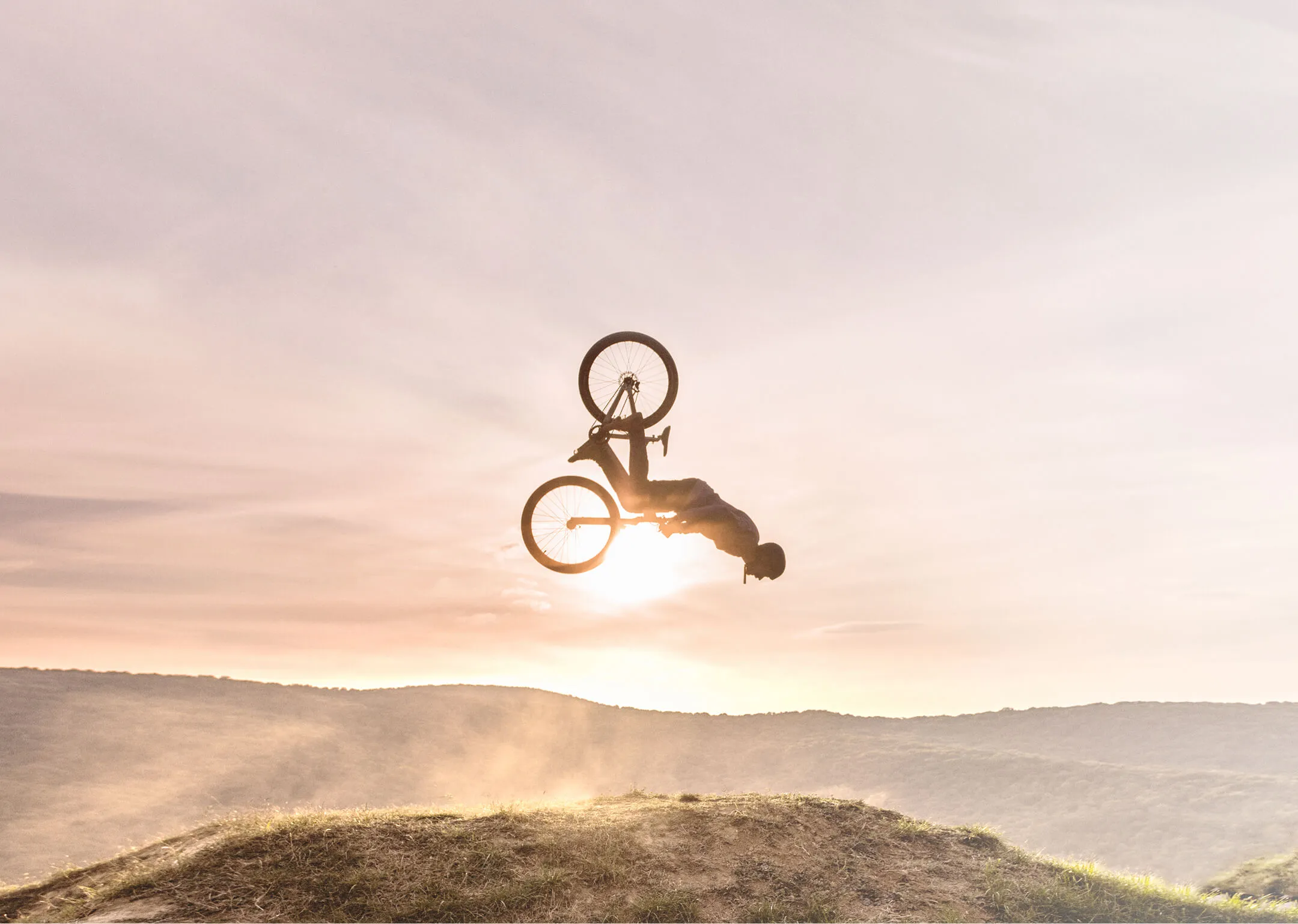 Mountainbiker in der Luft bei einem Rückwärtssalto über einer Hügelkante, im Hintergrund Sonnenuntergang mit nebliger Berglandschaft.