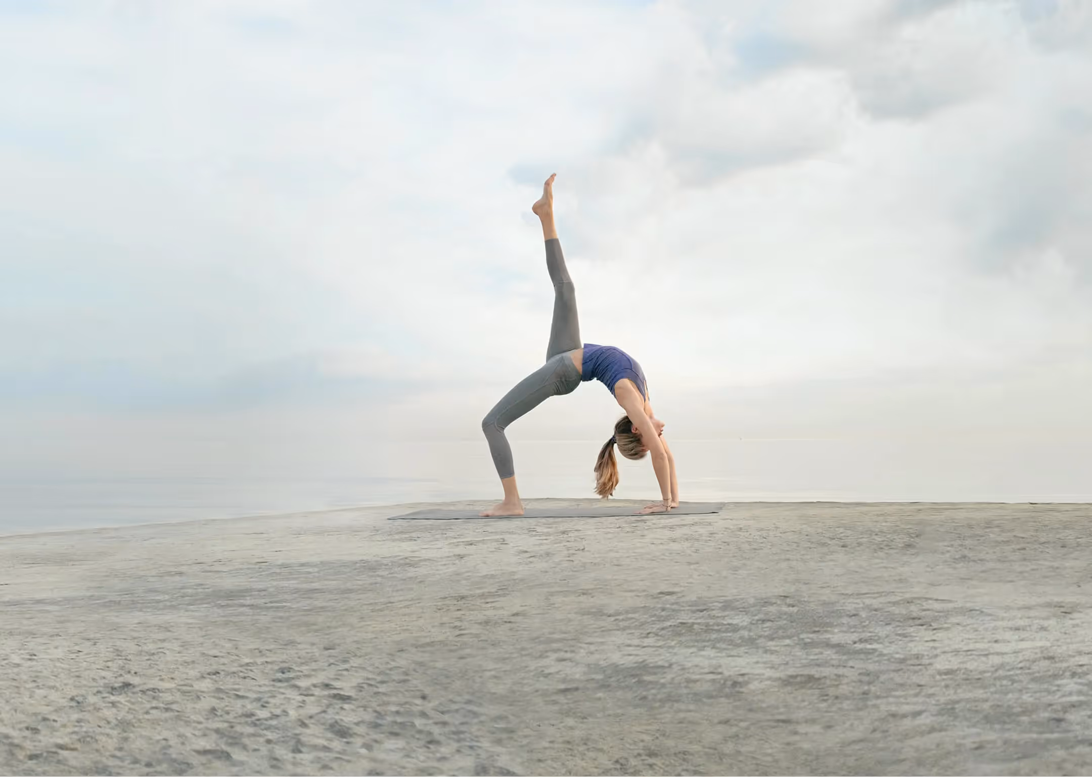 Frau macht Yoga auf einer weiten, flachen Steinfläche am Meer bei bewölktem Himmel. Sie befindet sich in einer Brücke mit einem Bein gerade nach oben gestreckt.