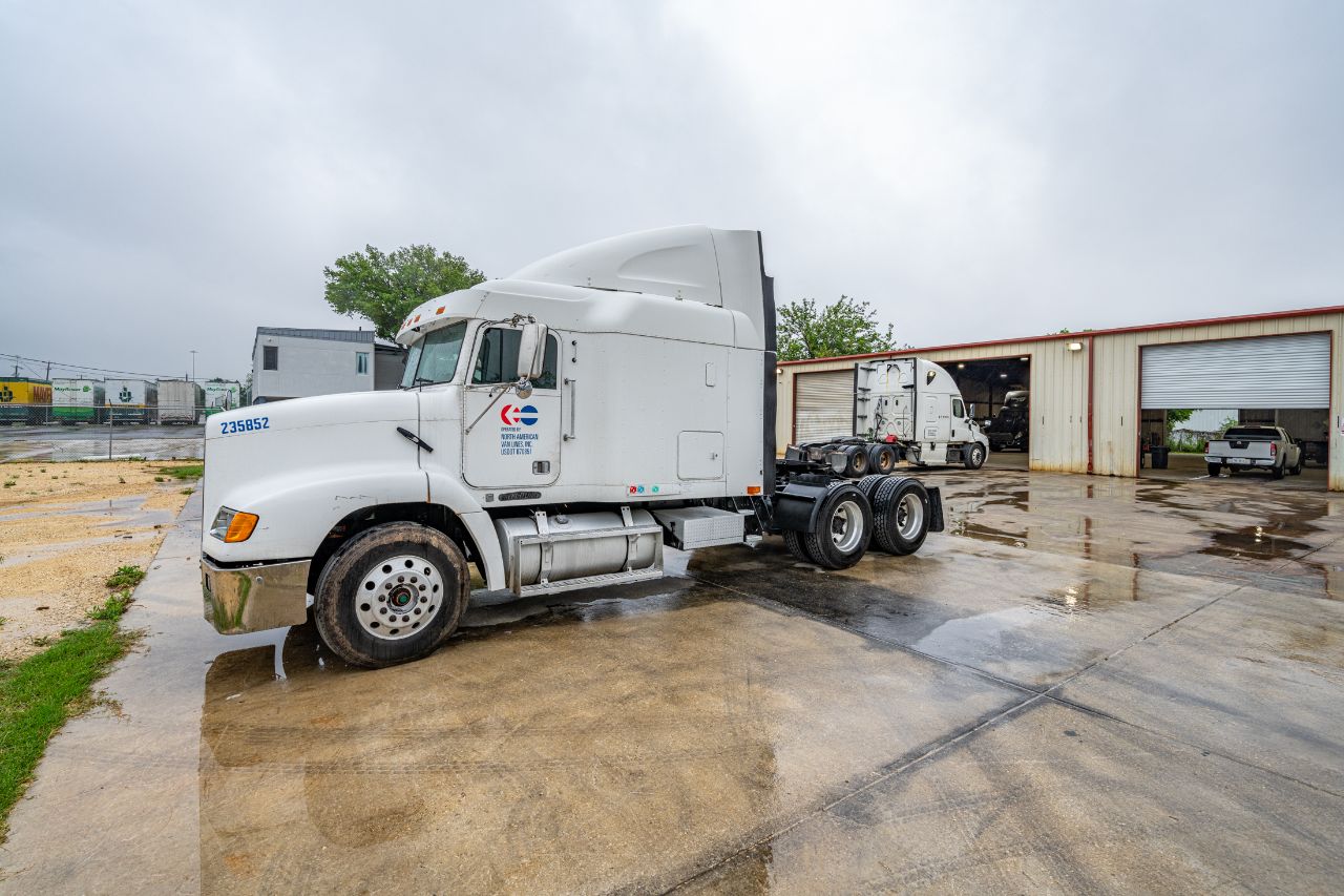 White semi-truck parked on wet concrete outside a diesel repair shop with open service bays on a cloudy, rainy day.