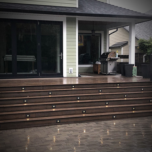 Outdoor wooden deck with built-in step lights and a BBQ grill under a covered patio.