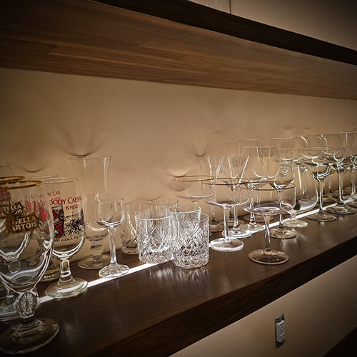 A wooden shelf with various clear glassware and some printed beer glasses illuminated by LED strip lighting underneath.