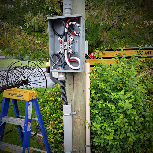 Electrical junction box mounted on a wooden utility pole with black and red cables inside, next to a blue ladder and green bushes.
