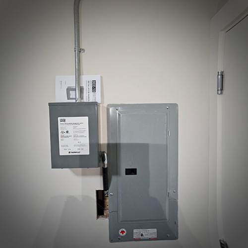 Two gray electrical panels mounted on a white wall beside a door, with conduit connecting the top panel and a shadow cast on the larger lower panel.