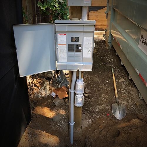 Outdoor electrical breaker panel with open door mounted on a wooden post beside a green dumpster, with a shovel resting on dirt ground.