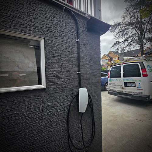 Electric vehicle charging station mounted on a gray textured exterior wall near a window, with a charging cable hanging below.