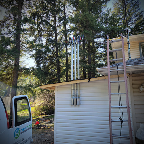 White house exterior with electrical conduit pipes mounted on the wall, a ladder leaning against the roof, and a van door open in a wooded area.
