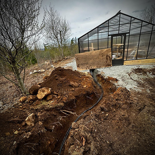 Trench with buried black cable leading to a greenhouse on a cloudy day, surrounded by trees and dirt piles.