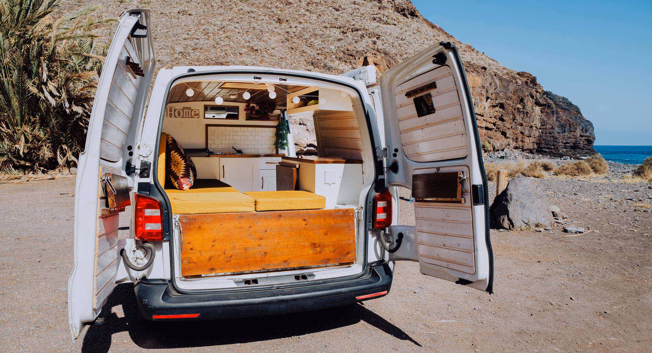 Parte trasera de la camper Majona con el portón abierto y el interior visible, junto a la costa.