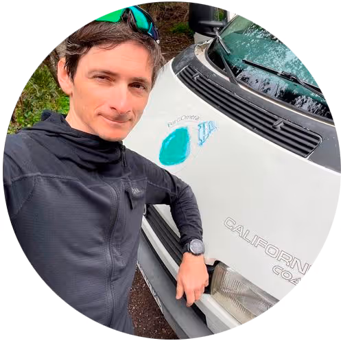 Hombre joven con chaqueta negra y gafas de sol verdes apoyado en la parte frontal de una furgoneta blanca con texto 'CALIFORNIA' visible.