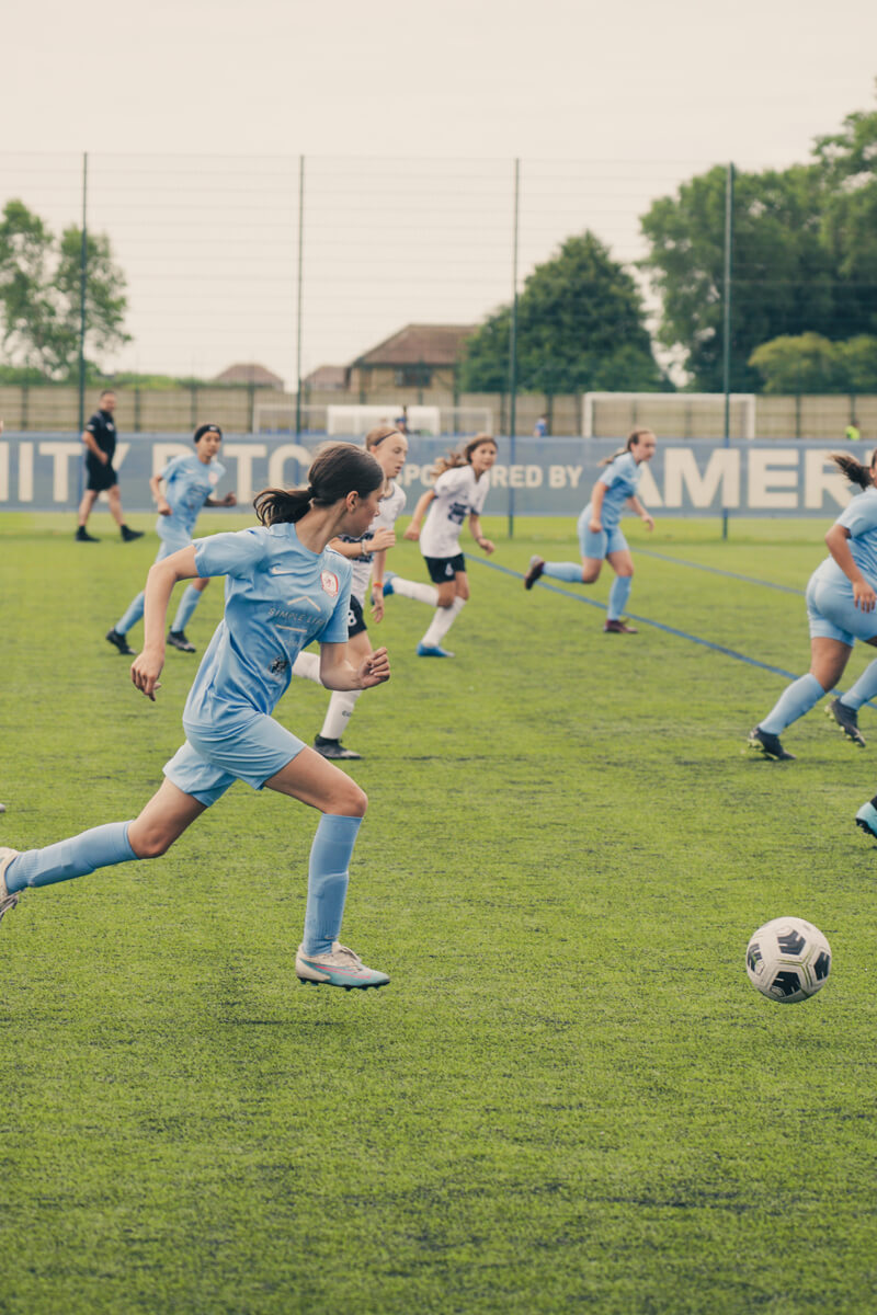 A female football player in a light blue kit sprinting with the ball during a match, with teammates and opponents in the background on an outdoor field.