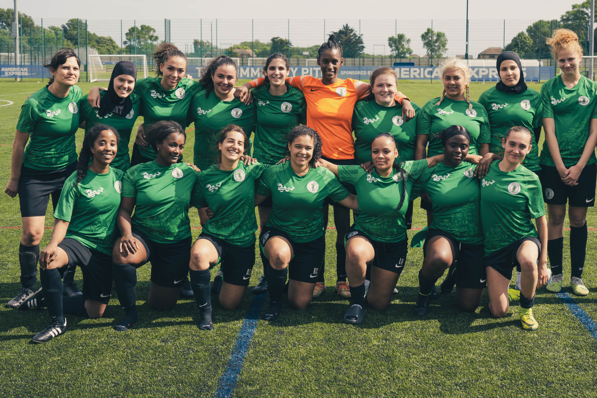 A women's football team poses on a pitch, wearing green shirts and black shorts. One player wears an orange goalkeeper jersey. They smile with arms around each other, with an "American Express" banner in the background.