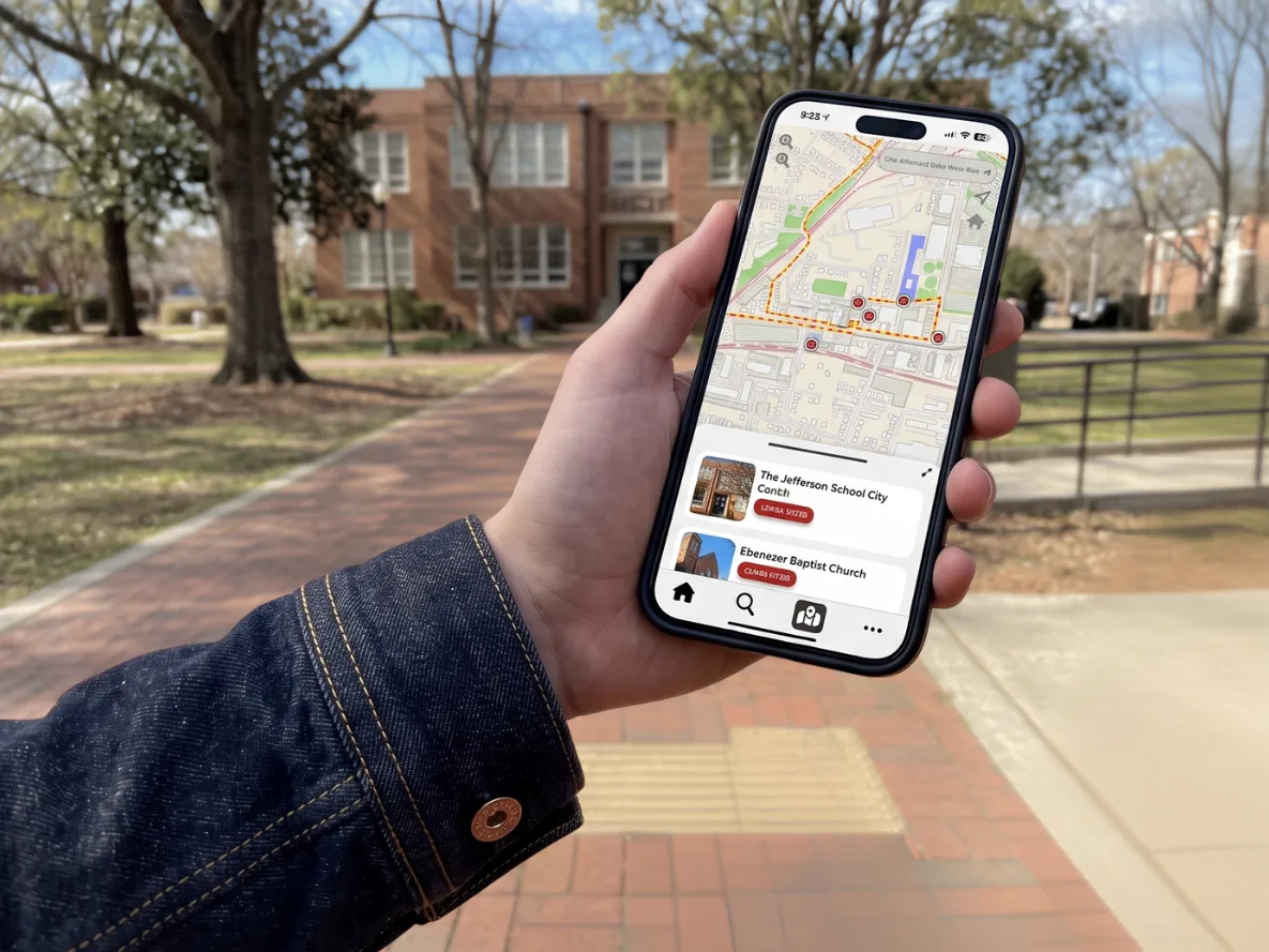 Hand holding smartphone showing a Guru map app with a route and location listings on a campus sidewalk.