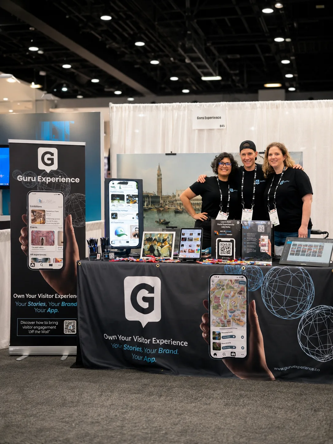 Dana, Suzie and Kealy wearing black Guru Experience shirts stand behind a branded booth with devices and displays.