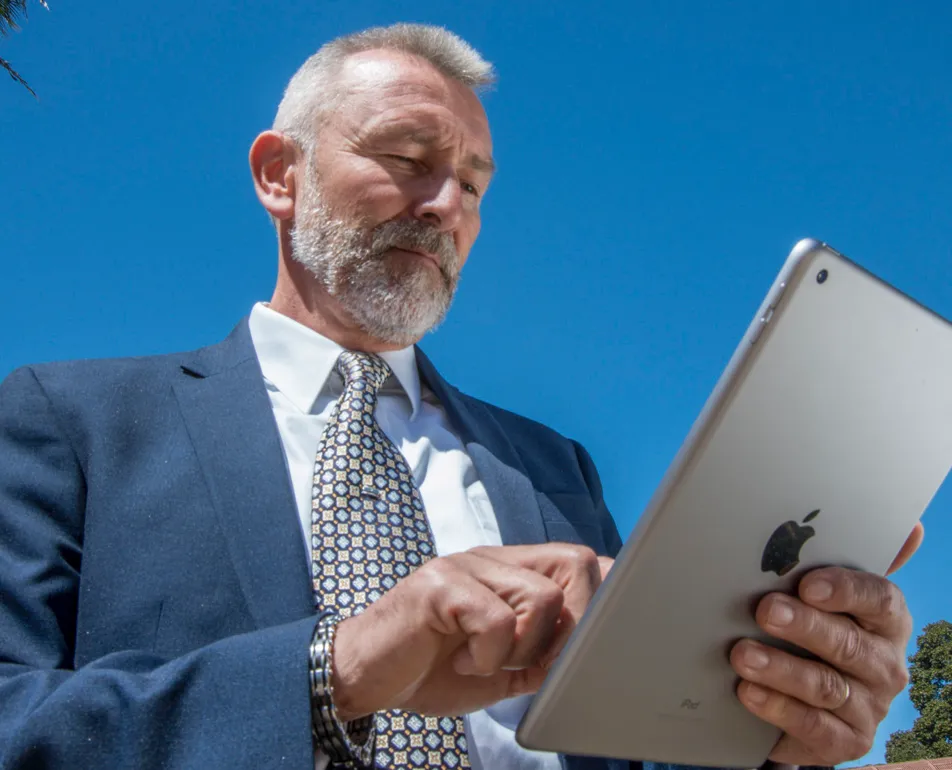 Man in a navy suit taps an iPad outdoors under a bright blue sky, the kind of mobile use the Balboa Park app supports across departments.