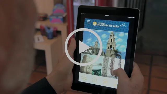 Video still with a play button: hands hold an iPad showing the San Diego Museum of Man app home screen with the California Tower.