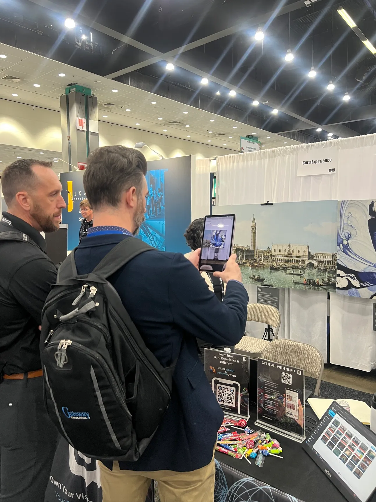 Two attendees at Guru's booth 845 aim a tablet at a large Venetian canal print, beside QR code signs and a laptop showing the CMS.