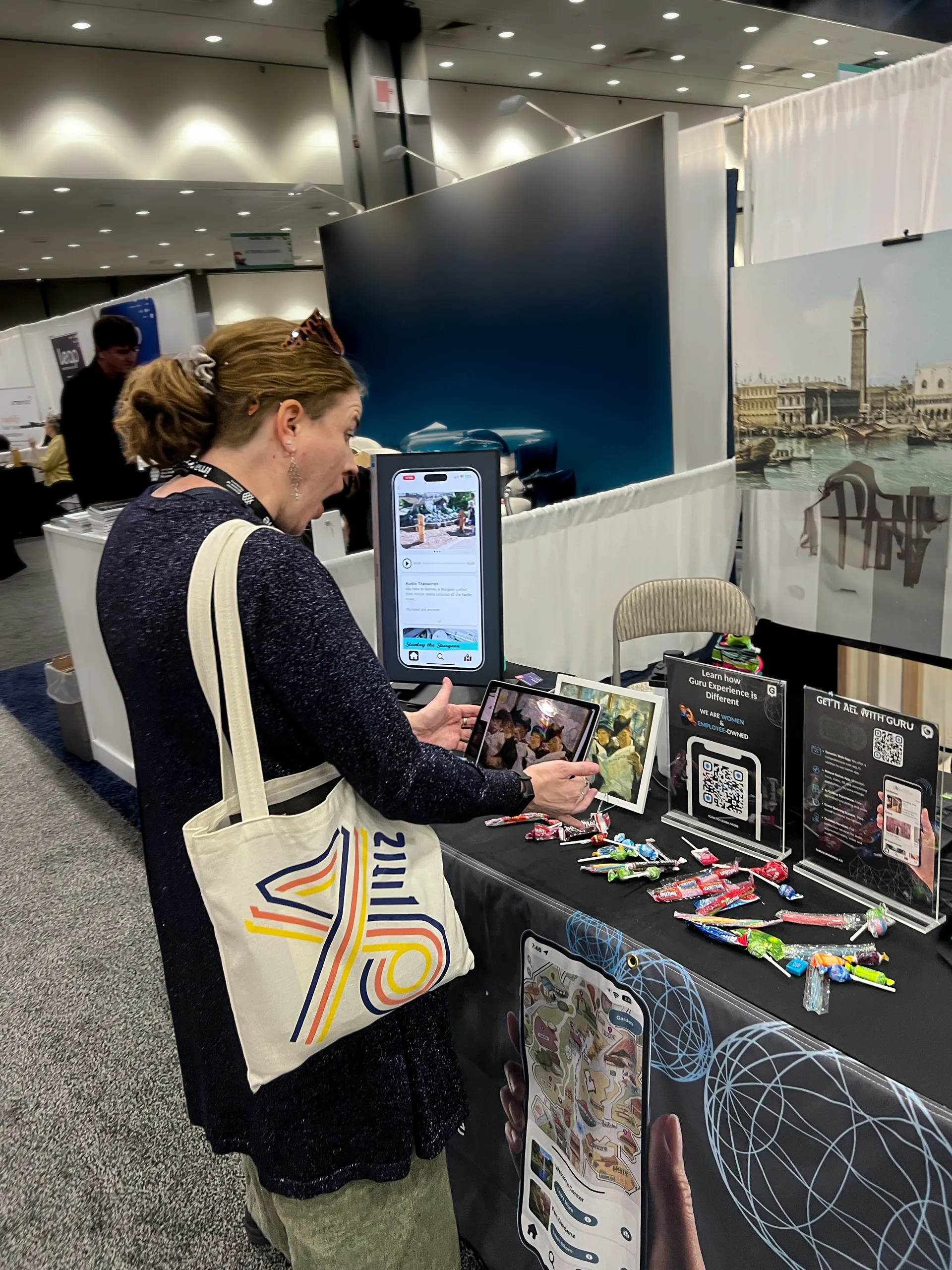 A conference attendee at Guru's AAM booth holds a tablet up to a framed painting to trigger an AR experience, beside QR code signs and phone demos.