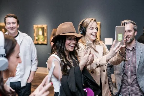 Visitors in a museum gallery wearing headphones and laughing while one films on a phone and another holds a microphone, artworks hanging behind them.