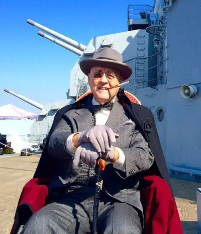 A costumed Franklin D. Roosevelt reenactor sits in a wheelchair on a battleship deck below the big gun turrets, cigarette holder in hand.