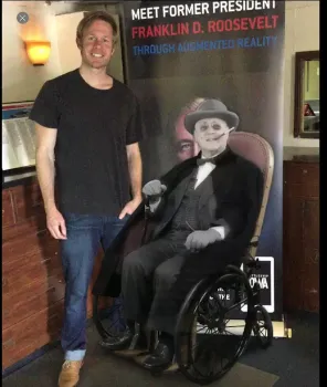 Visitor standing beside a banner reading Meet Former President Franklin D. Roosevelt Through Augmented Reality, with a seated FDR figure.