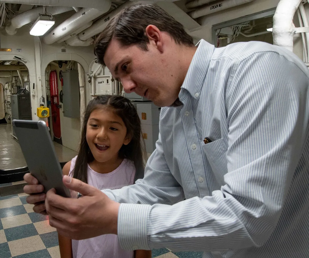 Inside a Battleship USS IOWA passageway, a man holds a tablet toward an exhibit while a wide-eyed girl watches the AR experience on the screen.