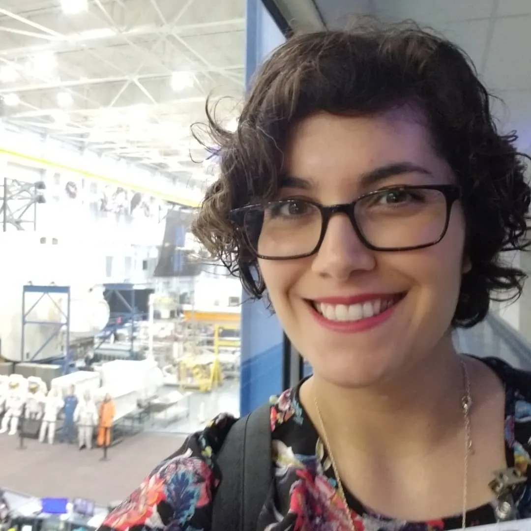 Guru's Director of Content Dana Duran smiles for a selfie at a window overlooking a hangar filled with spacecraft mockups and training equipment.