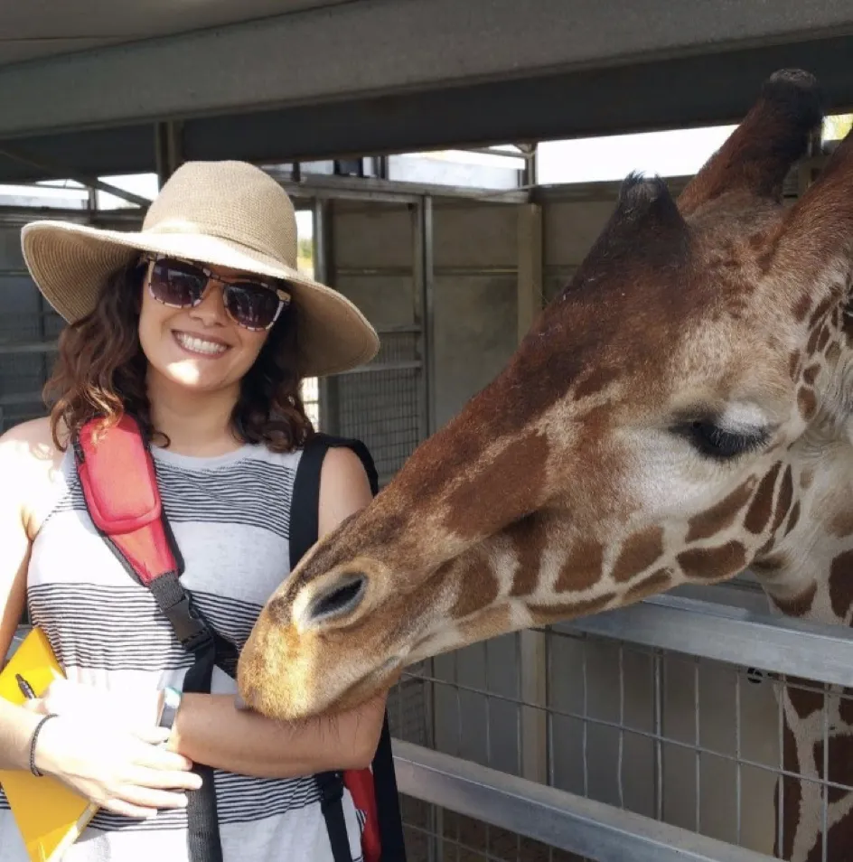 Dana Duran, Guru's lead storyteller, in a wide straw hat and sunglasses as a giraffe lowers its head toward her at a zoo fence rail.
