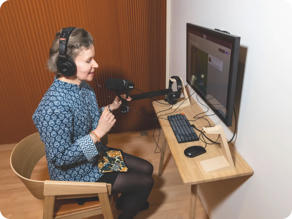 Visitor in headphones speaks into a boom microphone at a small recording desk with a wall-mounted screen, submitting her own story in the gallery.