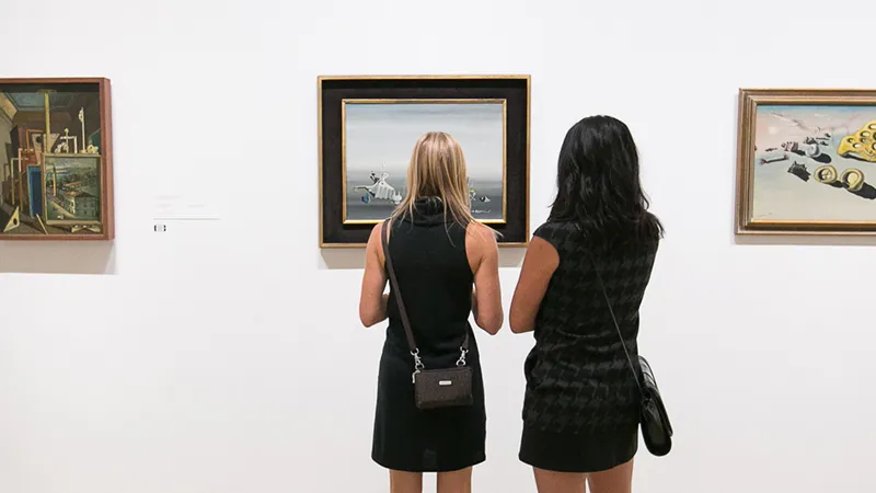 Two visitors in black stand shoulder to shoulder, seen from behind, studying framed surrealist paintings on a white gallery wall.