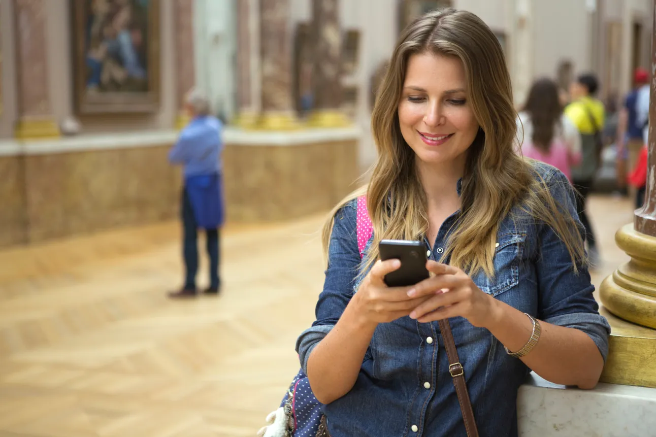Visitor in a denim shirt smiles at her phone while resting against a ledge in an ornate painting gallery, generating the app data the post describes.