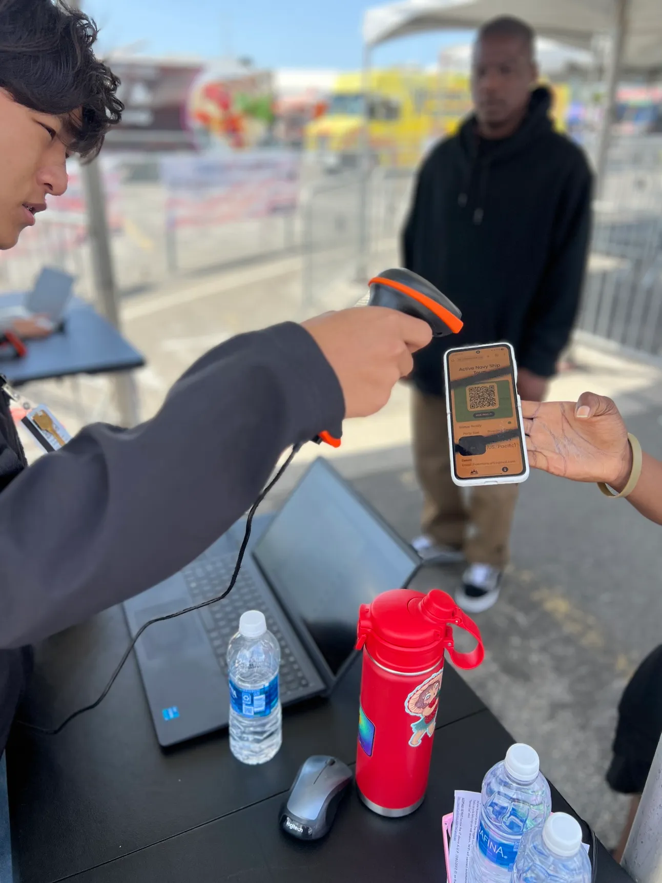 Event staffer uses a handheld scanner to read the QR boarding pass on a visitor's phone at an outdoor check-in table during LA Fleet Week.