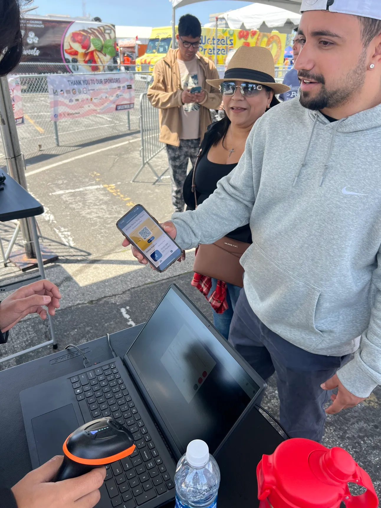 Visitor holds out a phone boarding pass to staff scanning barcodes at a laptop, with LA Fleet Week sponsor banners and food trucks behind.