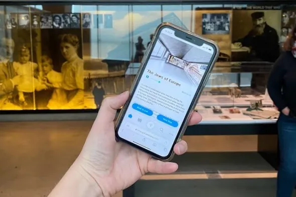 Hand holds a phone running the Holocaust Museum LA web guide, open to The Jews of Europe, in front of a gallery case of family photographs.