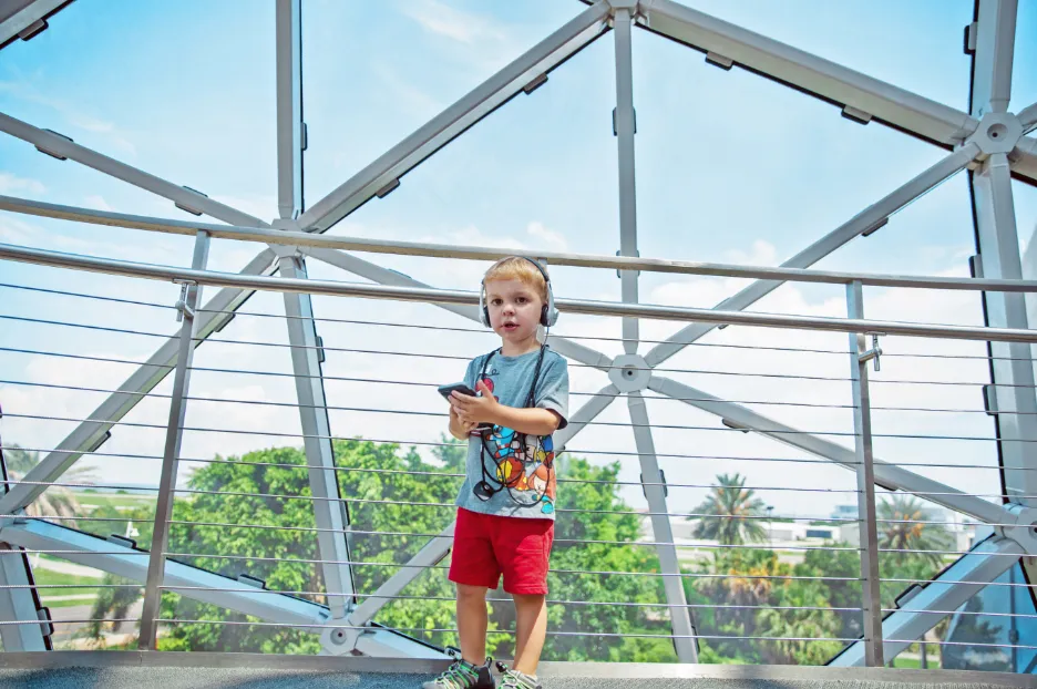 Young boy in headphones holds a phone on a museum's glass and steel walkway, palm trees visible outside, following the audio guide on his own.