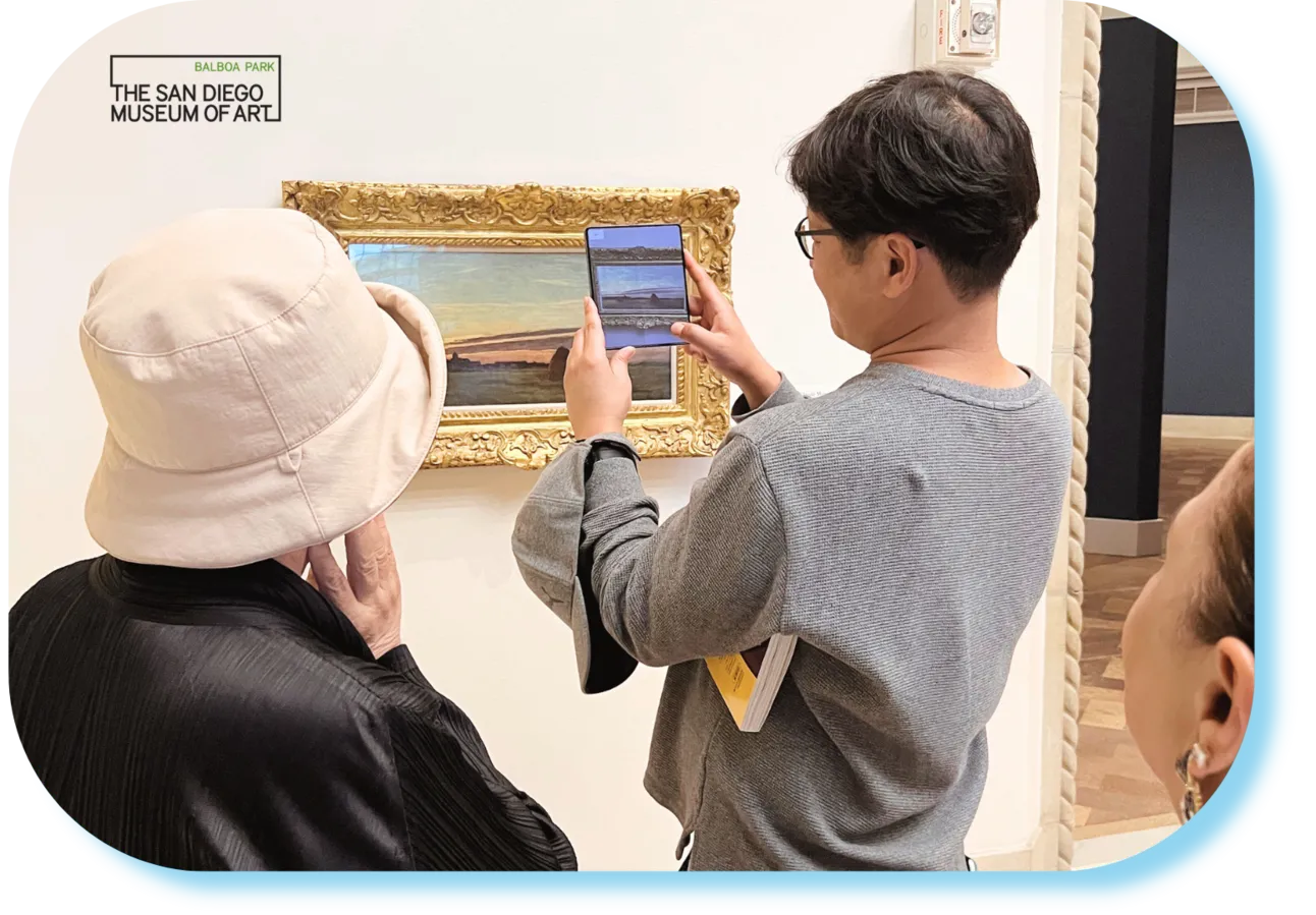 Two visitors at the San Diego Museum of Art hold a folding phone up to a gilt-framed seascape, the screen mirroring the painting for digital content.