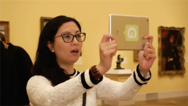 Visitor in a yellow painting gallery holds up a Guru-branded tablet and reacts with surprise as an augmented reality scene plays on screen.