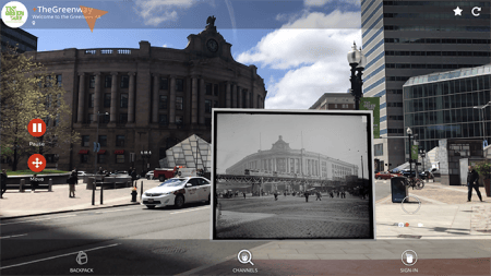 The Greenway app on a phone, holding a historical black and white photograph of Boston's South Station in front of the same street corner today.
