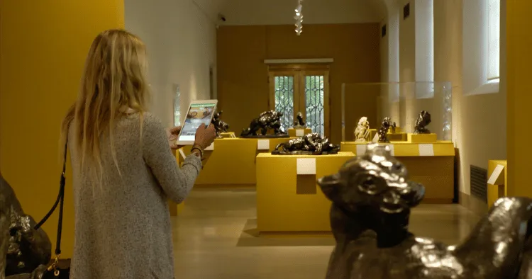 Museum visitor using a mobile digital guide while exploring animal sculptures displayed throughout a warmly lit gallery.