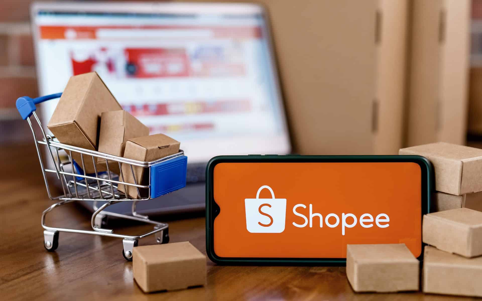 Notebook ligado com a pagina da plataforma shoppe. Um carrinho de supermercado miniatura com caixas dentro e um smatphone na horizontal com o logo da plataforma da Shoppe