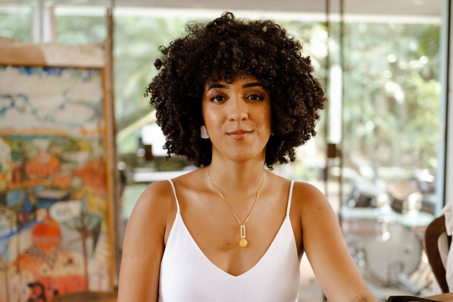 Fotografia da Nath Finanças, uma mulher preta de cabelo cacheado usando uma regata branca, colar dourado e brincos branco