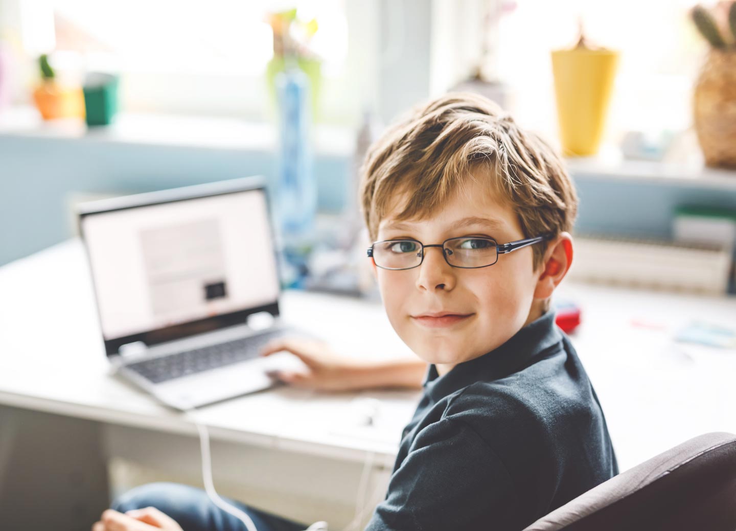 Young boy with glasses looks over his shoulder and smiles while on a laptop.