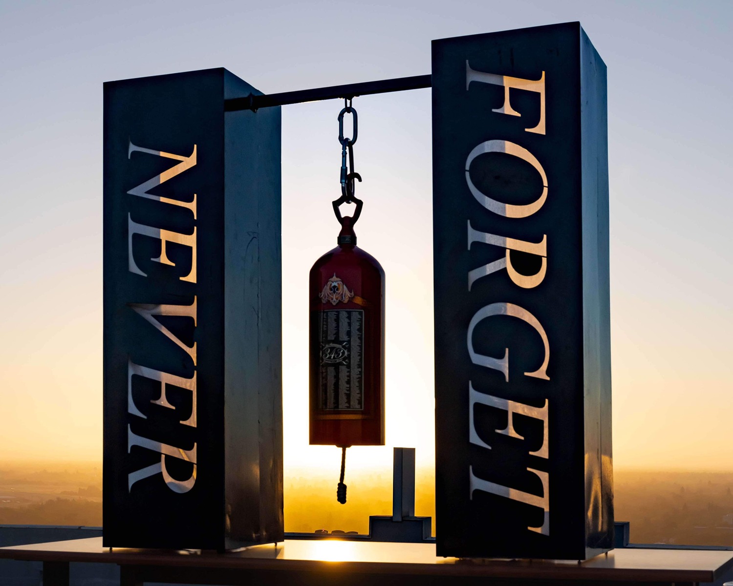 Metal sculpture with 'NEVER FORGET' text and a hanging fire extinguisher against sunset.
