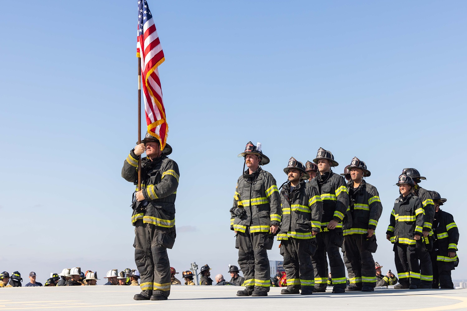 Group of firefighters in uniform standing outdoors, one holding an American flag on a pole.