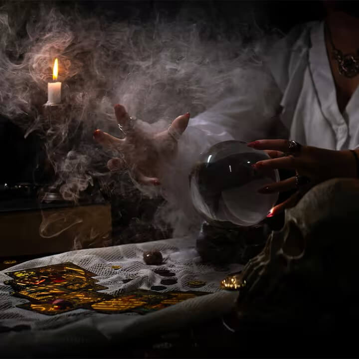 A hand with long red nails hovers over a crystal ball in a smoky room while cards lay strewn over the surface of a table