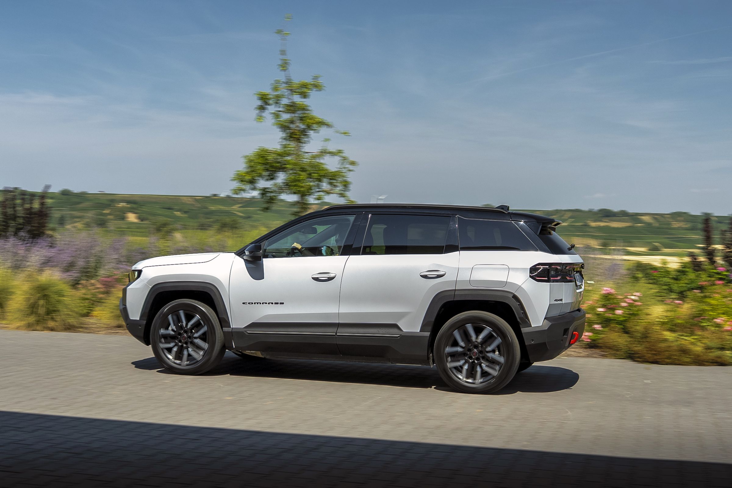 Neuer Jeep Compass 4xe Elektro in Weiß fährt seitlich an einer Blumenwiese vorbei