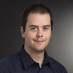 Garreth Tigwell is a white male with dark brown hair, smiling and wearing a dark blue shirt, while in front of a dark background.