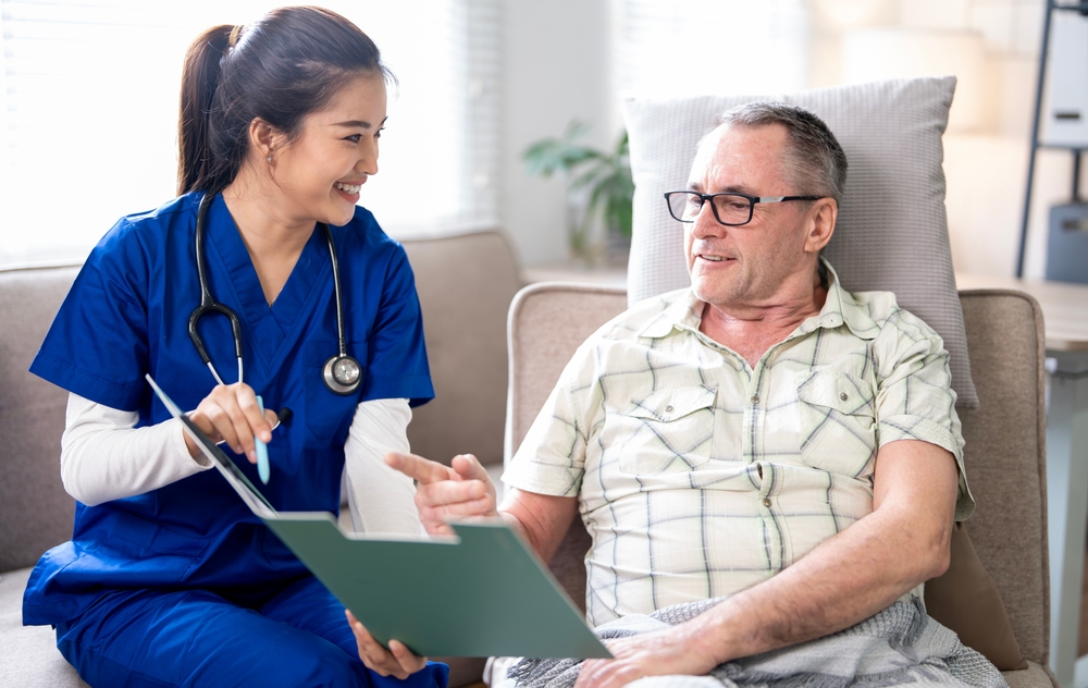 A nurse in blue scrubs with a stethoscope reviews a document with a senior man sitting on a couch.