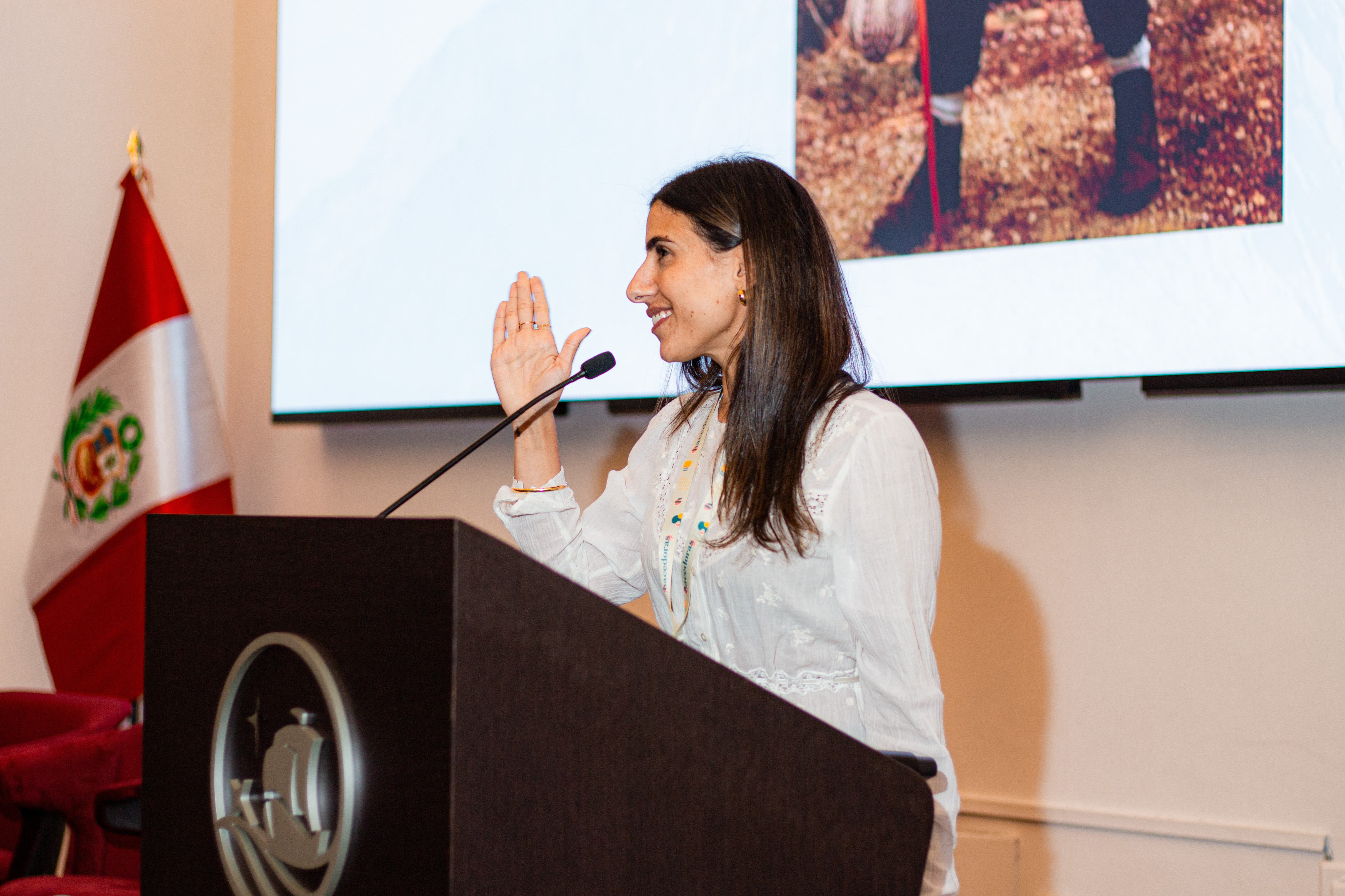Woman with long dark hair raises her hand speaking at a podium with Peru's flag in background.