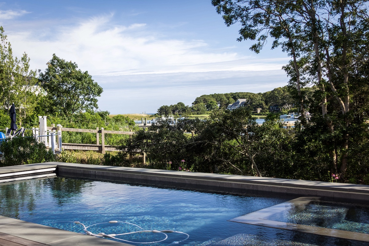 A beautiful pool in a luxury home's backyard abutting a body of water over some dune grass in Cape Cod MA